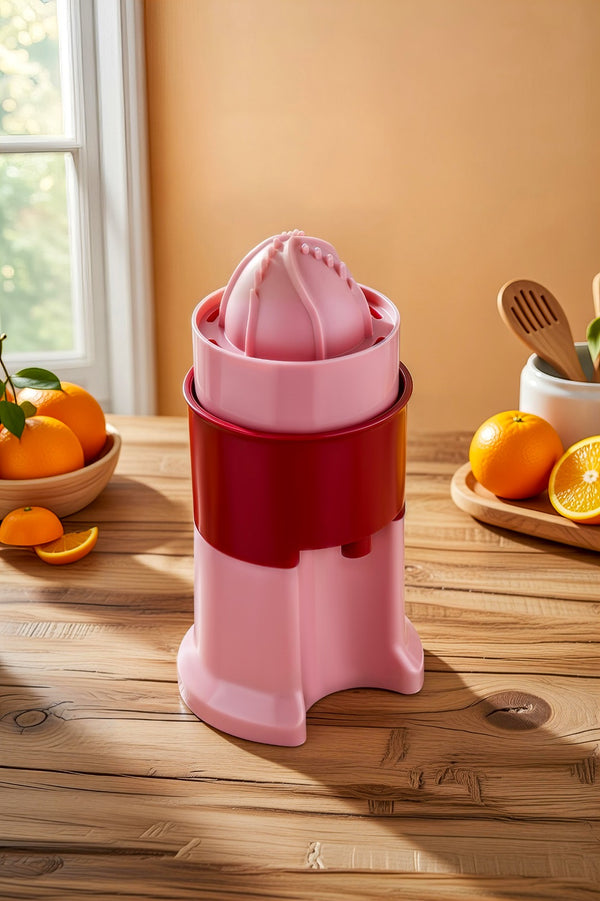 Pink manual juicer on a wooden table with oranges and kitchen utensils in the background.